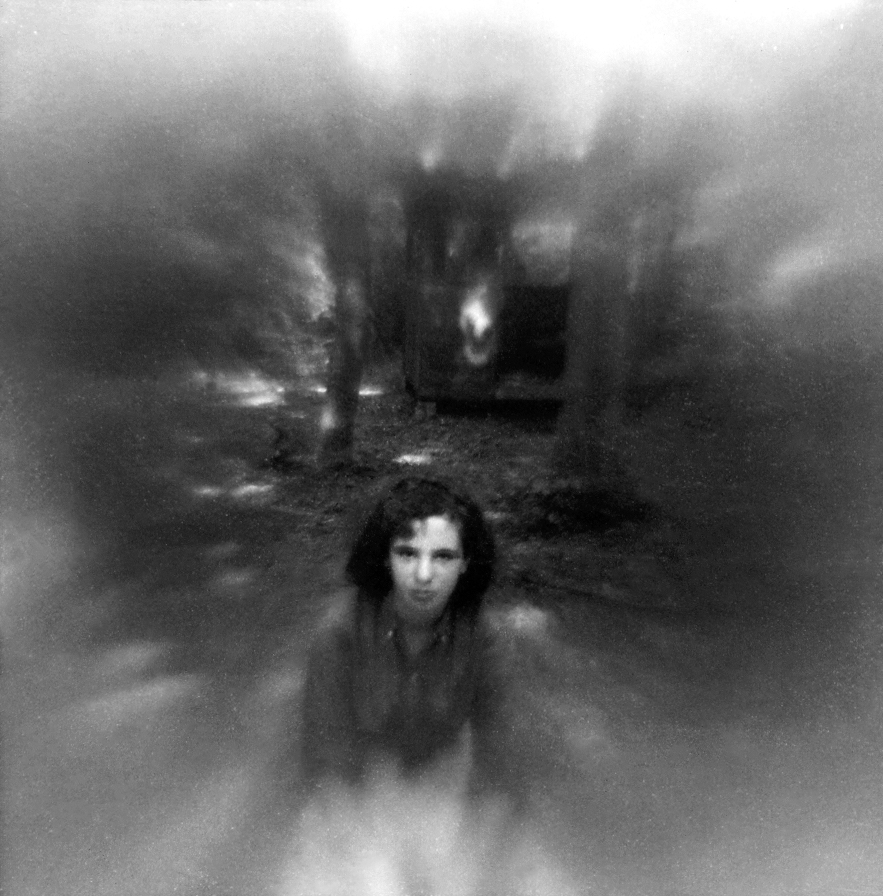 A grainy, atmospheric black and white photograph featuring a person&rsquo;s face centered at the bottom, surrounded by heavy blur and dark trees.