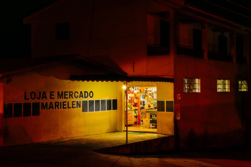 A small neighborhood shop called "Loja e Mercado Marielen" is lit warmly at night, with its open entrance revealing colorful products inside. The surrounding area is dimly lit, casting shadows on the building's exterior.