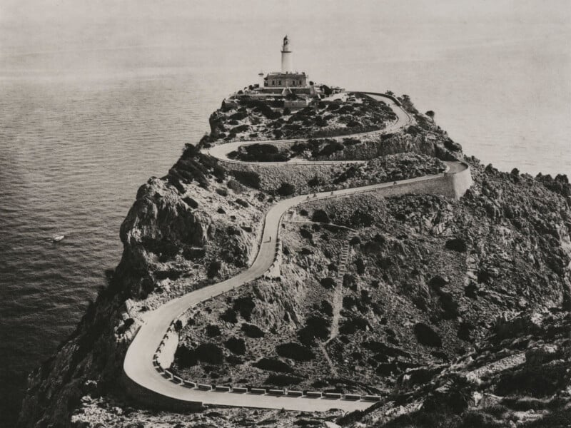 A winding road curves up a rocky cliff toward a lighthouse at the edge, surrounded by the sea; the landscape is dramatic and rugged, with sparse vegetation and waves visible below.