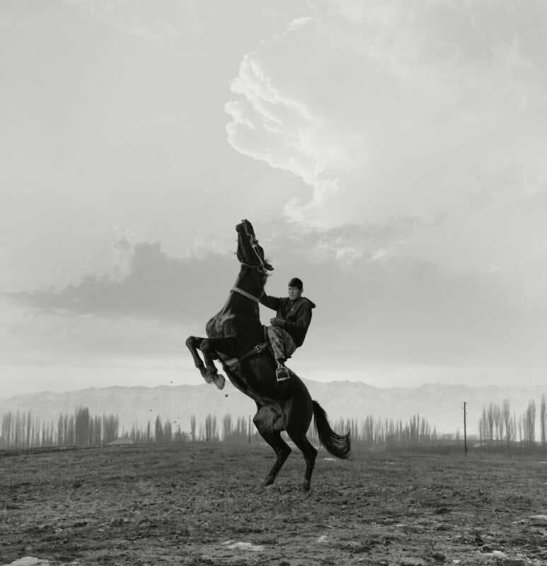 A person rides a rearing horse on a barren field with distant mountains and leafless trees under a dramatic cloudy sky. The image is in black and white.