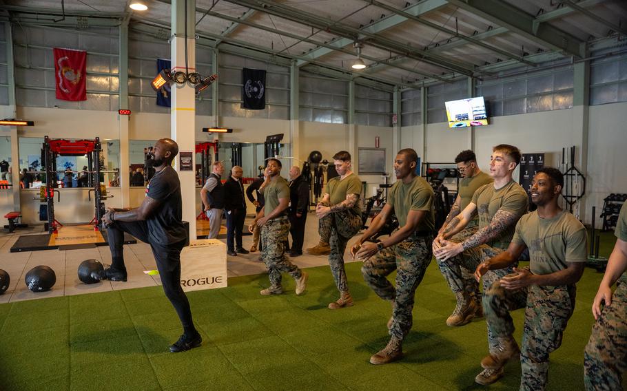 U.S. Marines follow a guest instructor through warm‑up movements during a group workout on artificial turf inside a fitness facility.