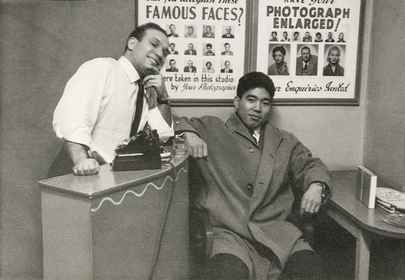 Two men pose in an office; one stands behind a counter with a typewriter, smiling, while the other sits beside him in a large coat. Posters behind them advertise photo enlargement and famous faces.
