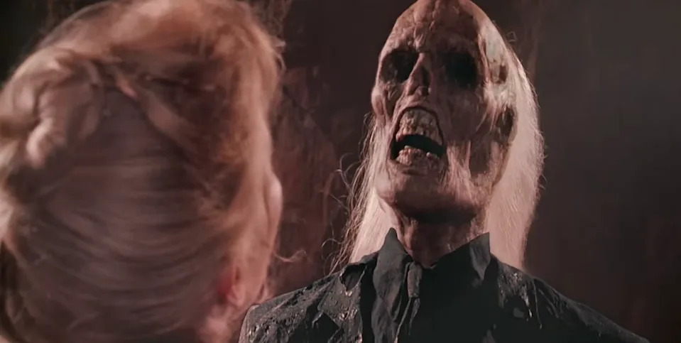A person faces a ghastly, skeletal figure with long, pale hair and dark clothing in a dramatic scene