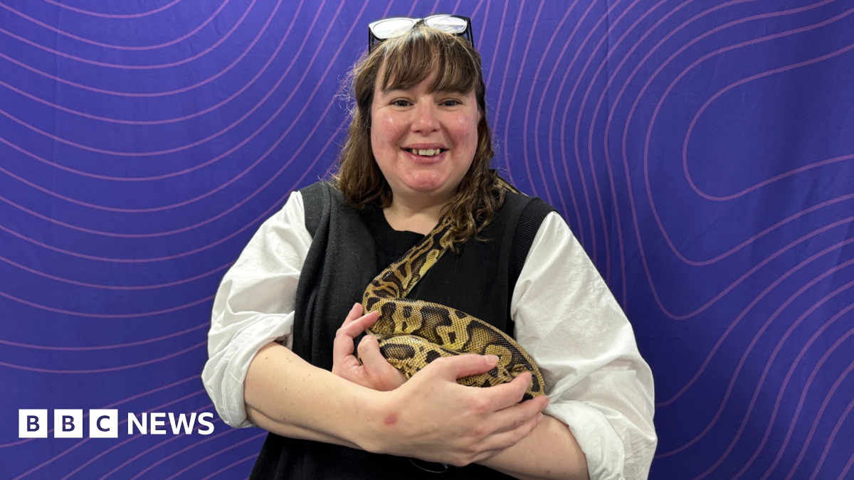 A woman, Vikki Buxton-Helyer, who is wearing glasses on top of her head and a white and black top, is holding a large yellow and black spotted snake in her arms. She is smiling and standing in front of a purple background.