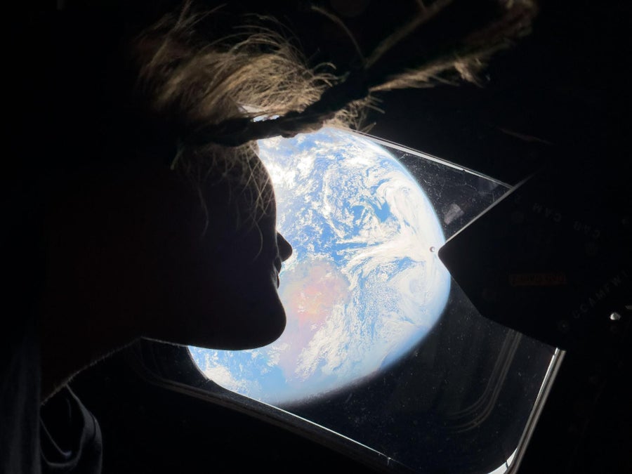 Astronaut Christina Koch looks out a window of the Orion spacecraft with a pale-white earth large in the distance.