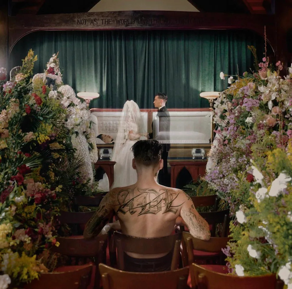 The cover art for Yeat's 'ADL' album. The art depicts a shirtless man with tattoos sitting in a chapel, facing a coffin and a couple in wedding attire. Surrounded by floral arrangements.