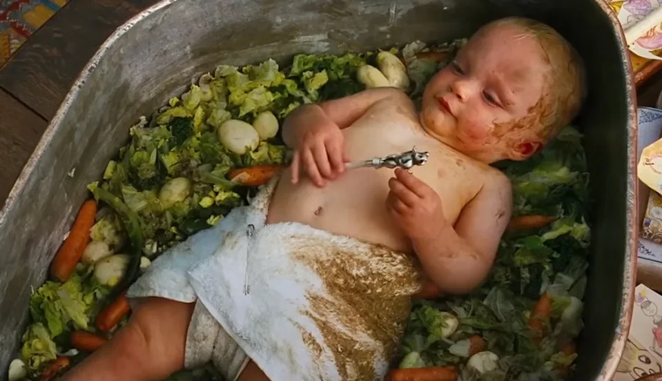 Baby laying in a tub of vegetables, playing with a utensil, spoofing a cooking pot scene