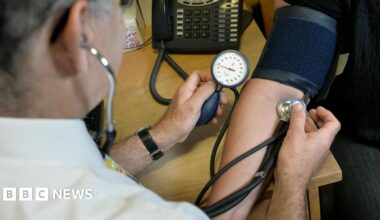 A doctor conducting a blood pressure test on a patient. He is holding a medical instrument against an arm whilst monitoring a gauge. The patient has a dark blue wrap around their arm.