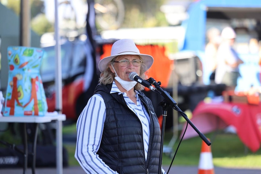 a woman in a striped top and vest speaks into a microphone and wears a hat.