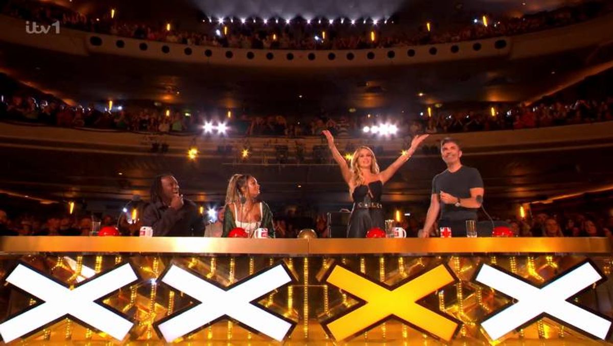 A group of individuals are gathered on a stage, raising their hands in celebration. The stage is adorned with a golden barrier featuring large white and yellow letters spelling out "XXX". Spotlights are positioned overhead, illuminating the scene.