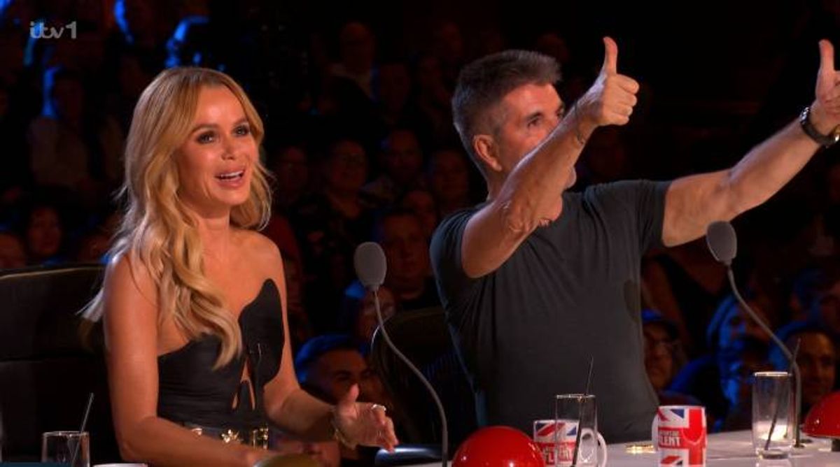 An individual with long, light-colored hair, dressed in a black sleeveless top, stands at a microphone, gesturing with a thumbs-up. Beside them, another individual in a black t-shirt makes a similar gesture, both in a brightly lit setting with a blurred audience in the background.