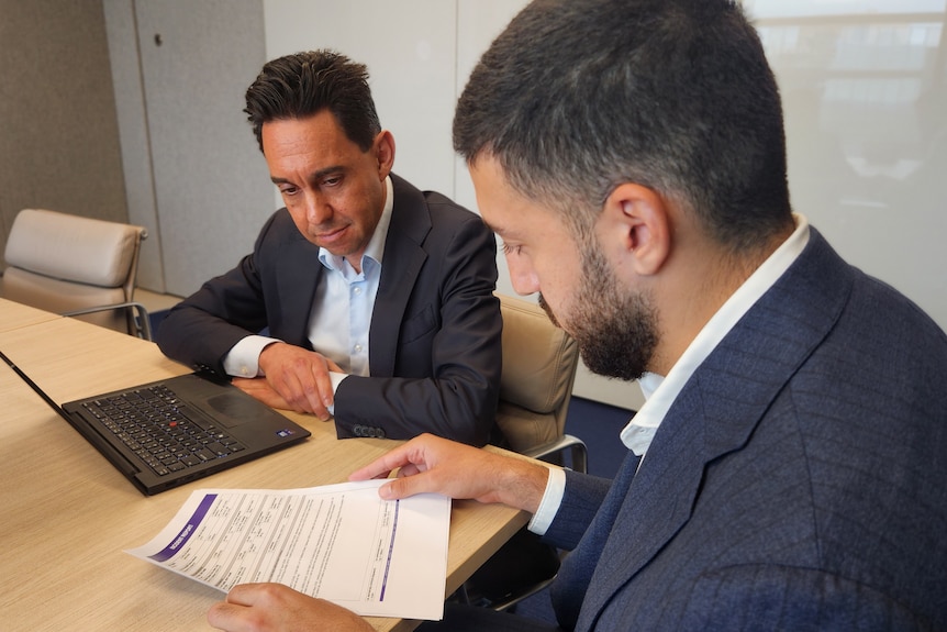 Two men sit at a table looking at a document, with a laptop open in front of them.