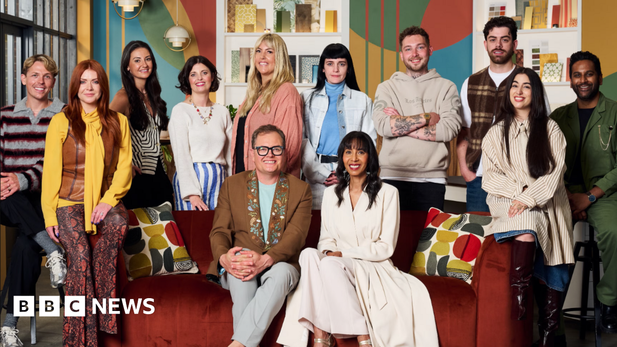 Alan Carr is sat down on a red settee wearing a brown jacket and grey trousers. He is sat next to judge Michelle Ogundehin, who is dressed in a cream jacket and matching skirt. They are joined by ten people, who are all contestants on the series Interior Design Masters. Sophie from Stoke on Trent is among the contestants and is second left in the photograph. She has long red hair and is wearing a yellow shirt, patterned red trousers and a brown gilet. Ben, from Kinver, is stood behind the red sofa with his arms folded, wearing a beige hoodie.