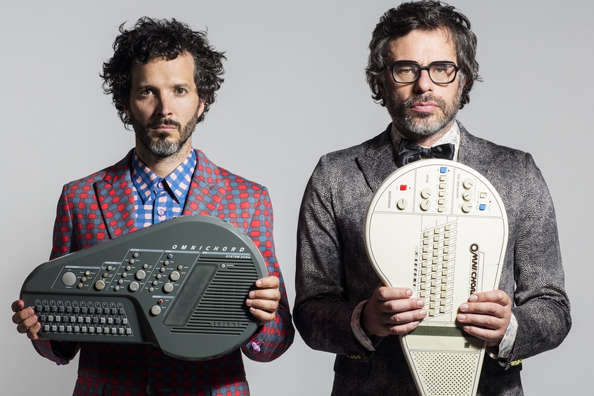 Bret, wears red and grey polka dot suit and Jemaine grey suit, black tie, both, serious, stand holding an instrument.