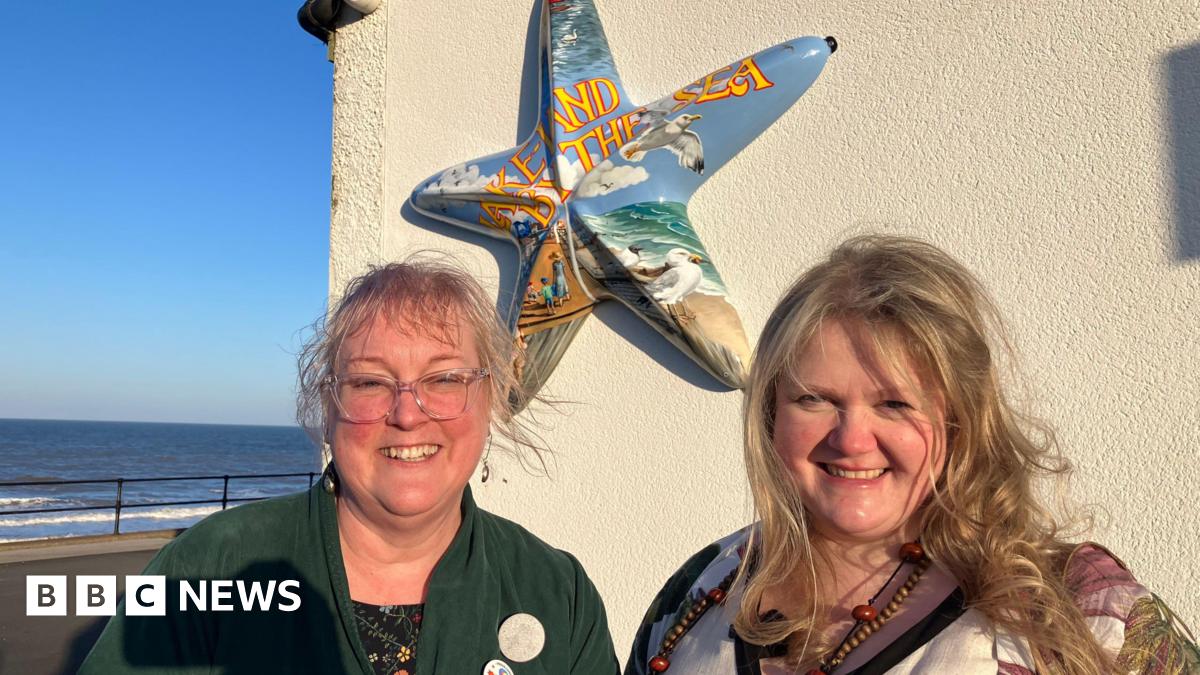 A woman with a blonde fringe and hair tied back is wearing large clear-rimmed glasses and a green cardigan and is smiling into the camera. Next to her is a woman with long blonde hair also smiling into the camera. Behind them is a wall with a starfish sculpture which says 'Lake-Land By The Sea' in yellow lettering with a background of the seaside with seagulls, sky, sand and the sea. The actual sea can be seen behind them.