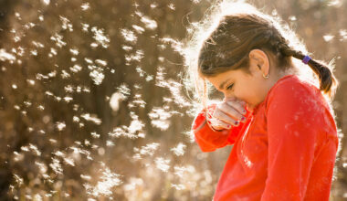 A young girl standing outside is surrounded by floating plant fluff and pollen. She's sneezing.
