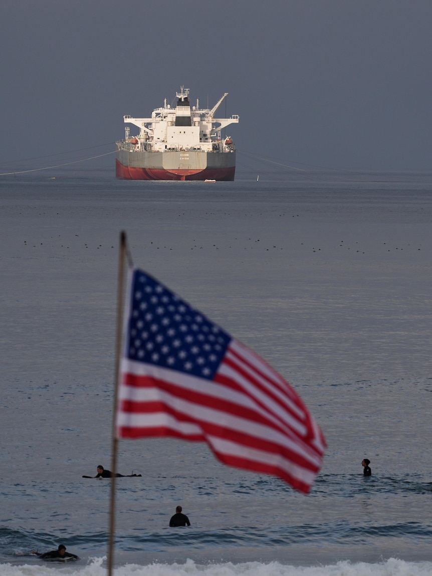 Surfers wait for waves at the beach, with a crude oil tanker in the background and a US flag in the foreground