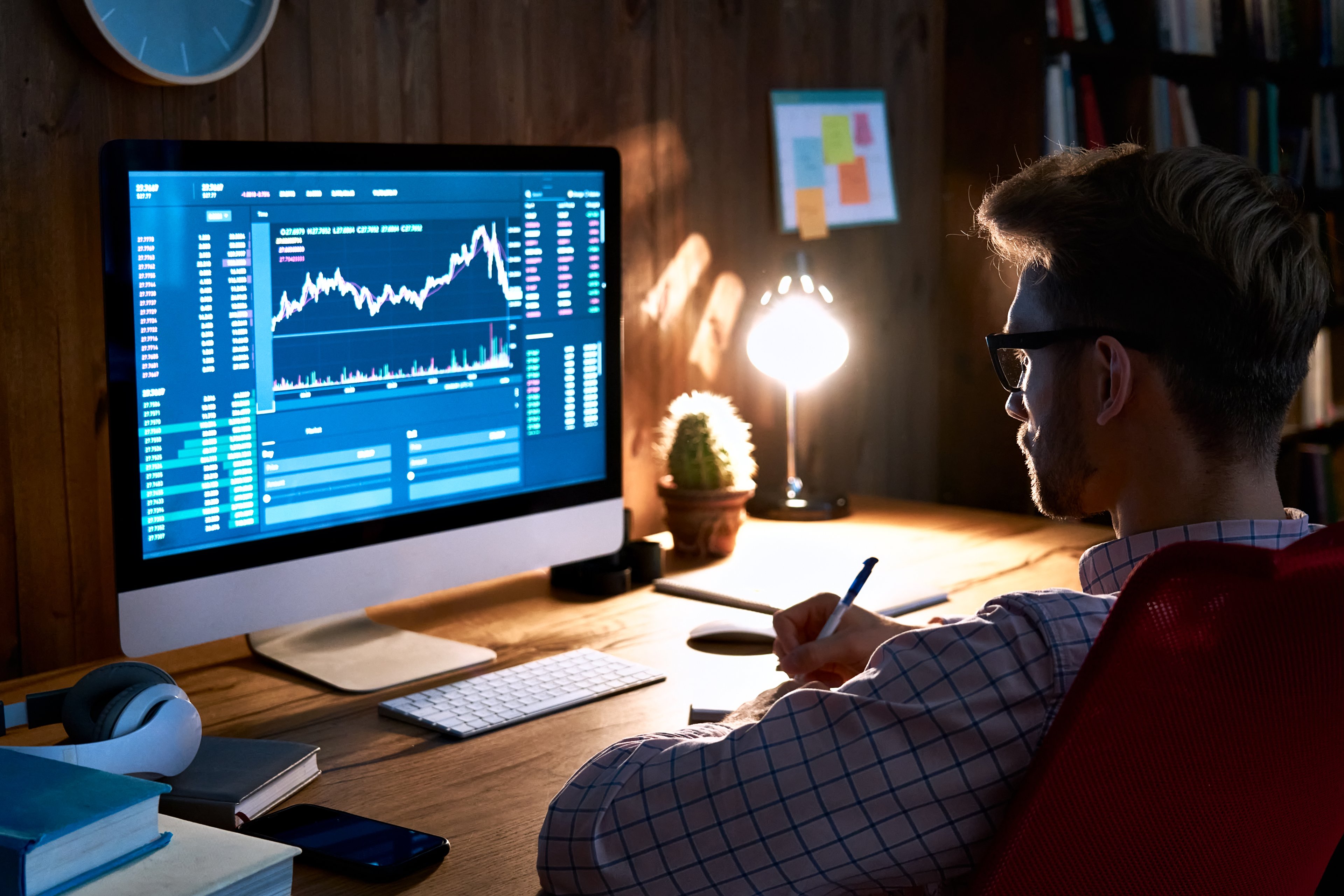 An investor sitting in a dark room looking at their computer screen, which is displaying a stock or cryptocurrency chart.