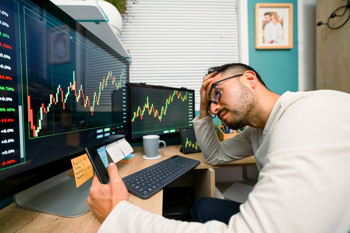 A worried-looking person holds their forehead while sitting at a desk with two monitors showing stock charts.