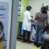 A patient care room is photographed from outside the room. A poster with Spanish language is visible, as is a doctor in a white coat, caring for a child on an exam table. 