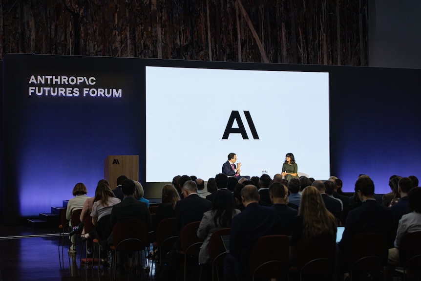 A man and a woman sit on a stage at a forum with a crowd underneath.