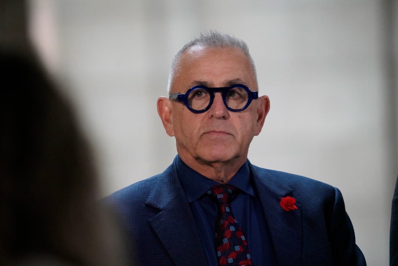 A man in a dark blue blazer and patterned red tie speaks with media in a government building.