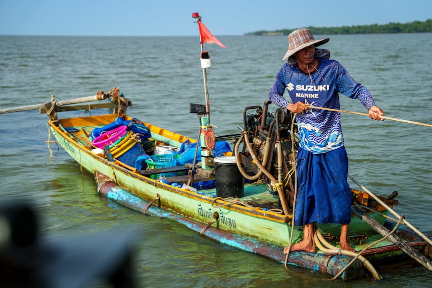 A man on a small yellow fishing dingy stands at the rear of the boat, pulling himself to shore with a rope.