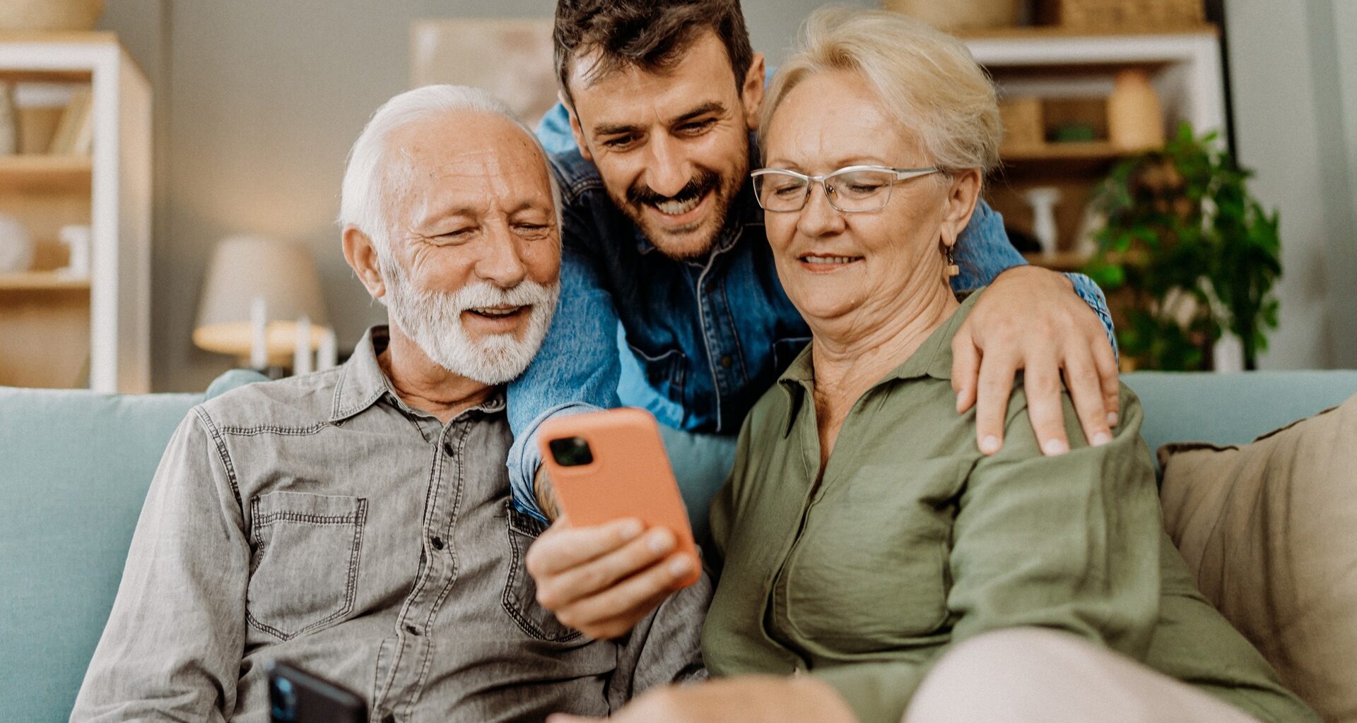 Younger Man leaning over his older parents sitting on the couch while they show pictures on a phone
