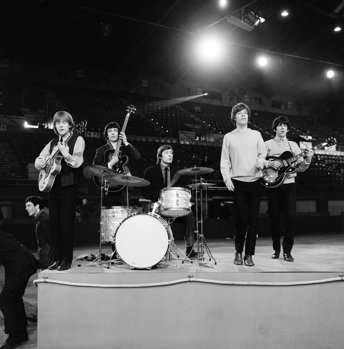 The Rolling Stones rehearse on a stage under lights in 1964