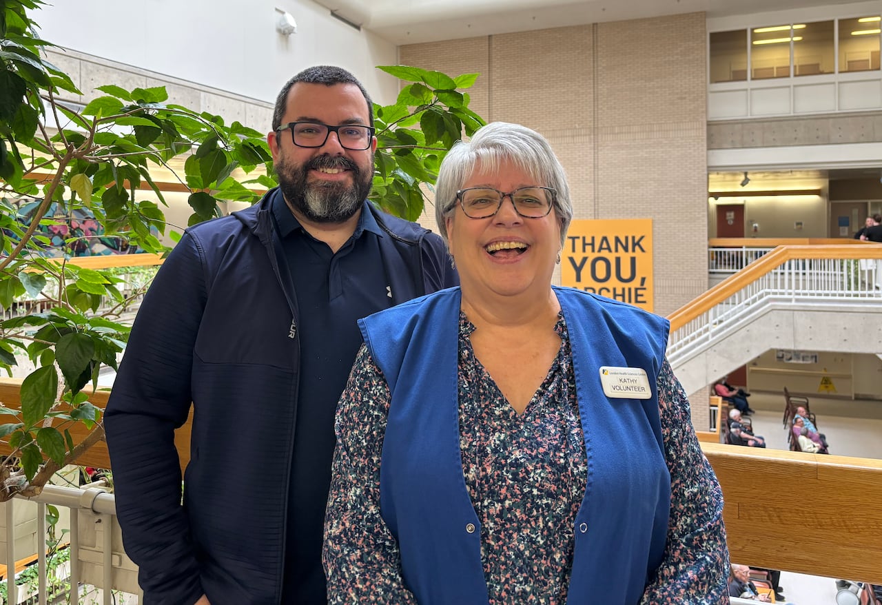 A man and a woman stand smiling in a hospital atrium. The woman wears a blue volunteer vest and a name tag that reads “Kathy.”
