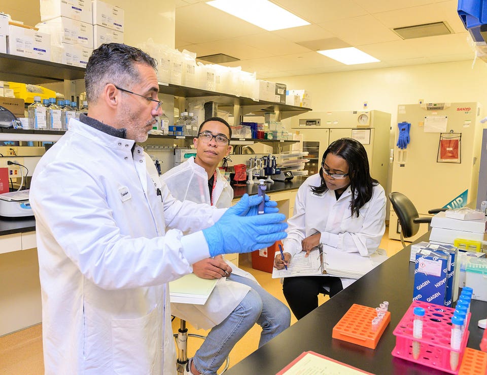 Three people in white lab coats; two take notes