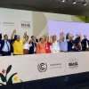 A group of representatives from multiple countries, wearing formal attire, lifts their joined hands. They stand behind a table that reads "COP30, Brazil".