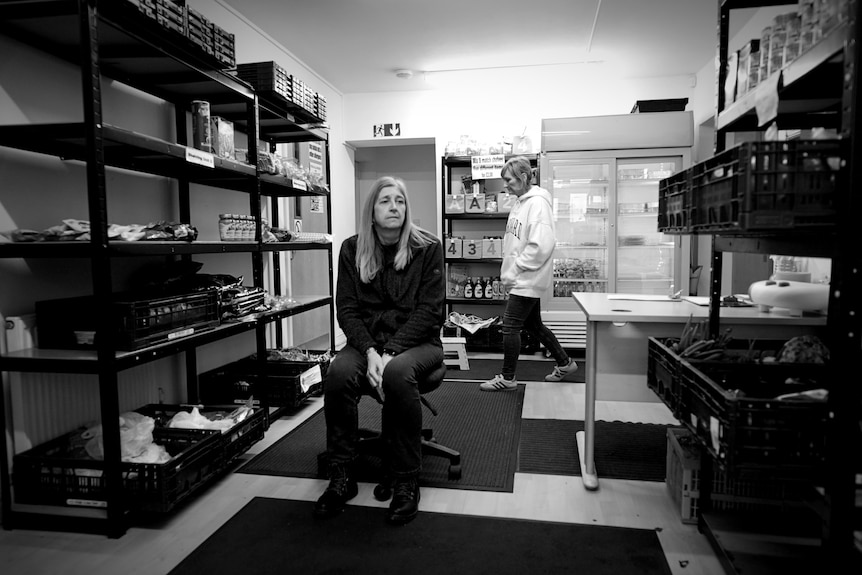 A woman sits on an office chair at a food bank as another woman walks past behind her.