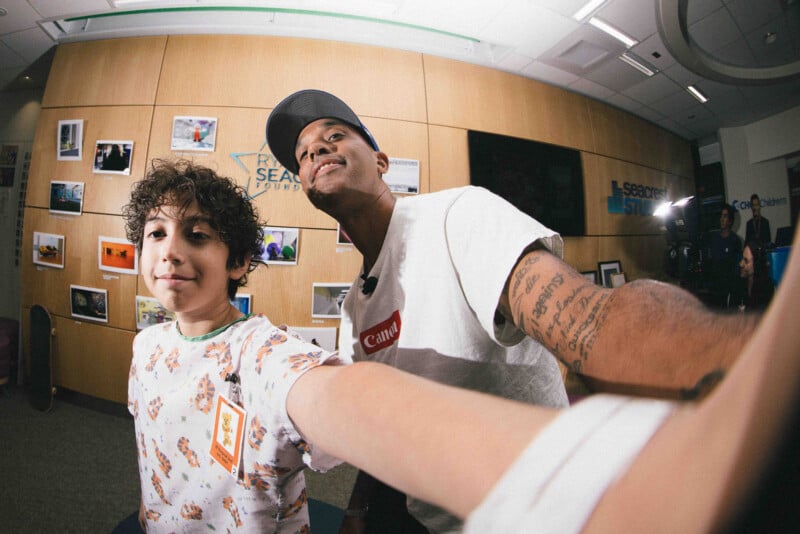 A young boy and a man take a selfie together in a brightly lit room with wooden walls decorated with photos and signs. The man has tattoos on his arm and wears a cap, while the boy smiles at the camera.