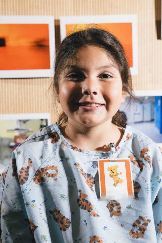 A smiling child wearing a hospital gown with tiger prints and a badge featuring a cartoon bear, standing in front of a wall with colorful photos.
