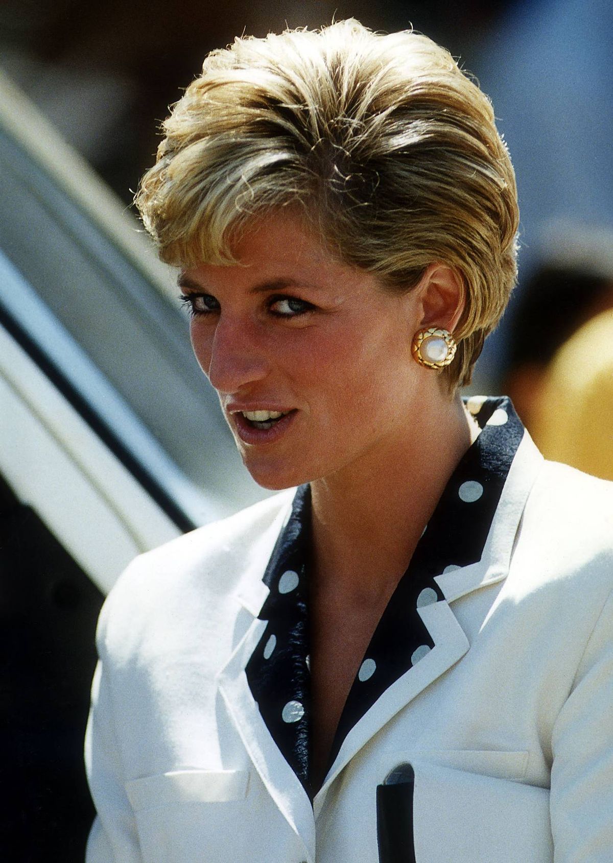 A close-up image of an individual with short, light-colored hair and a refined appearance, possibly engaged in an outdoor event. The individual is wearing a white blouse and has a pair of pearl earrings, exuding a sense of elegance and poise.