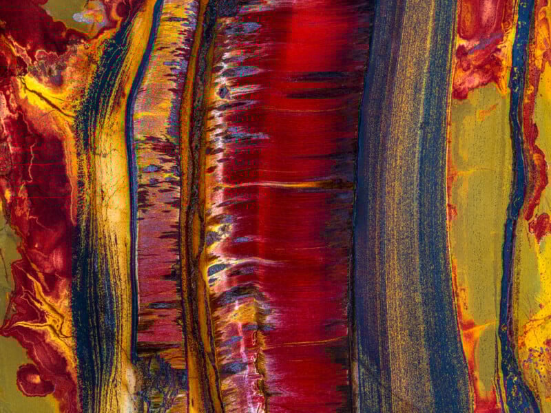 Abstract painting with bold vertical streaks of red, orange, gold, and dark blue, blending and overlapping with textured, fluid edges and vibrant, fiery color contrasts.