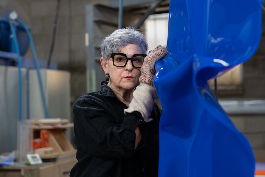 A woman with short greying hair holds a crumbled blue sheet of plastic upright.
