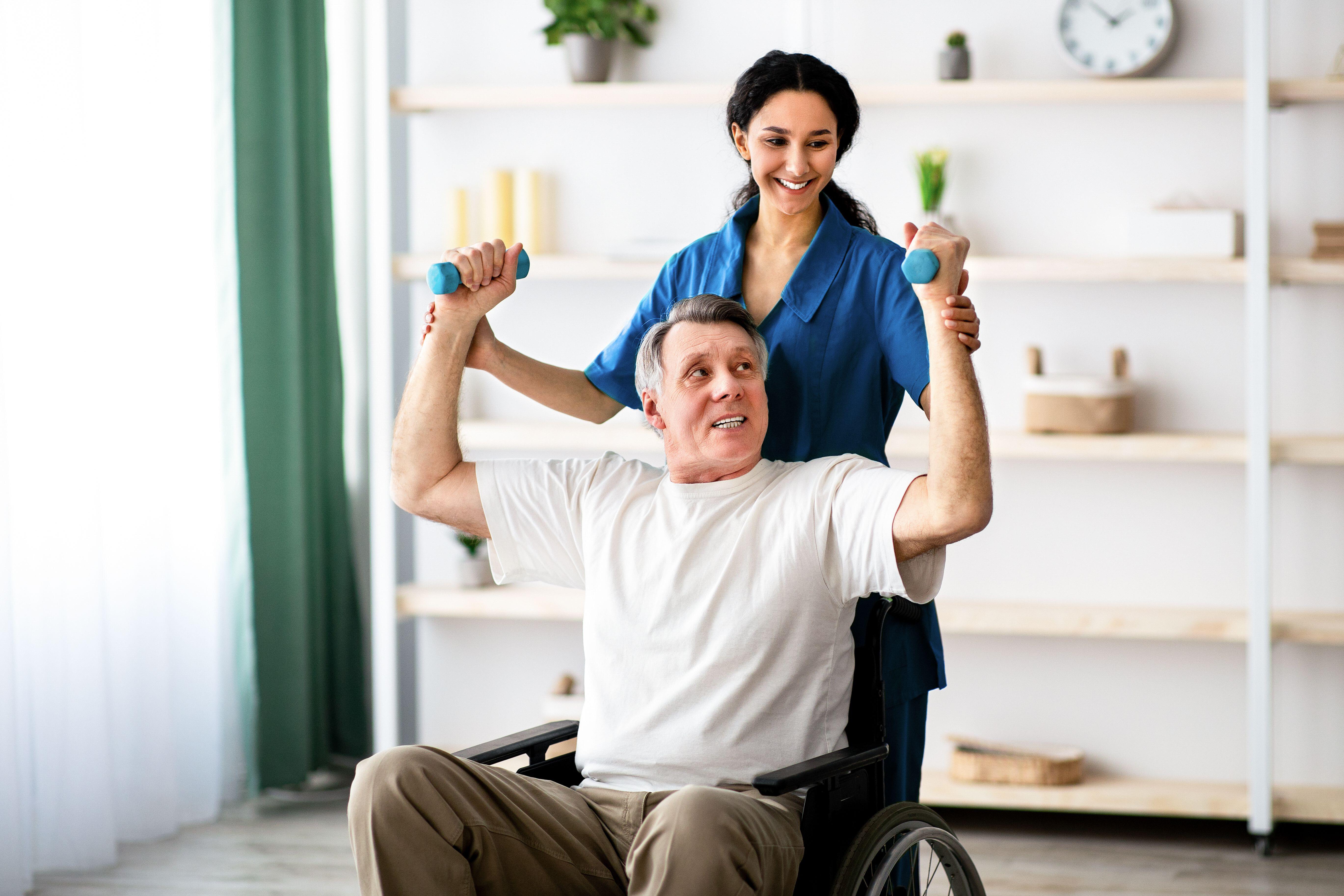 A female physiotherapist standing behind a man sitting in a wheelchair and helping him to lift small dumbells in each of his hands