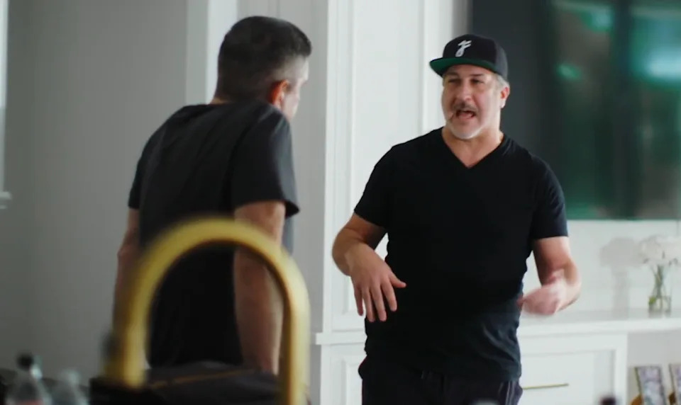 Two men are casually talking inside a room. One man is wearing a black cap and t-shirt, gesturing with his hands