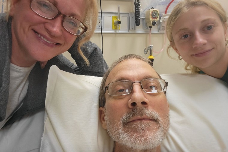 Eric Tennant lays in a hospital bed as Becky, left, and Amiya, right lean in for a selfie