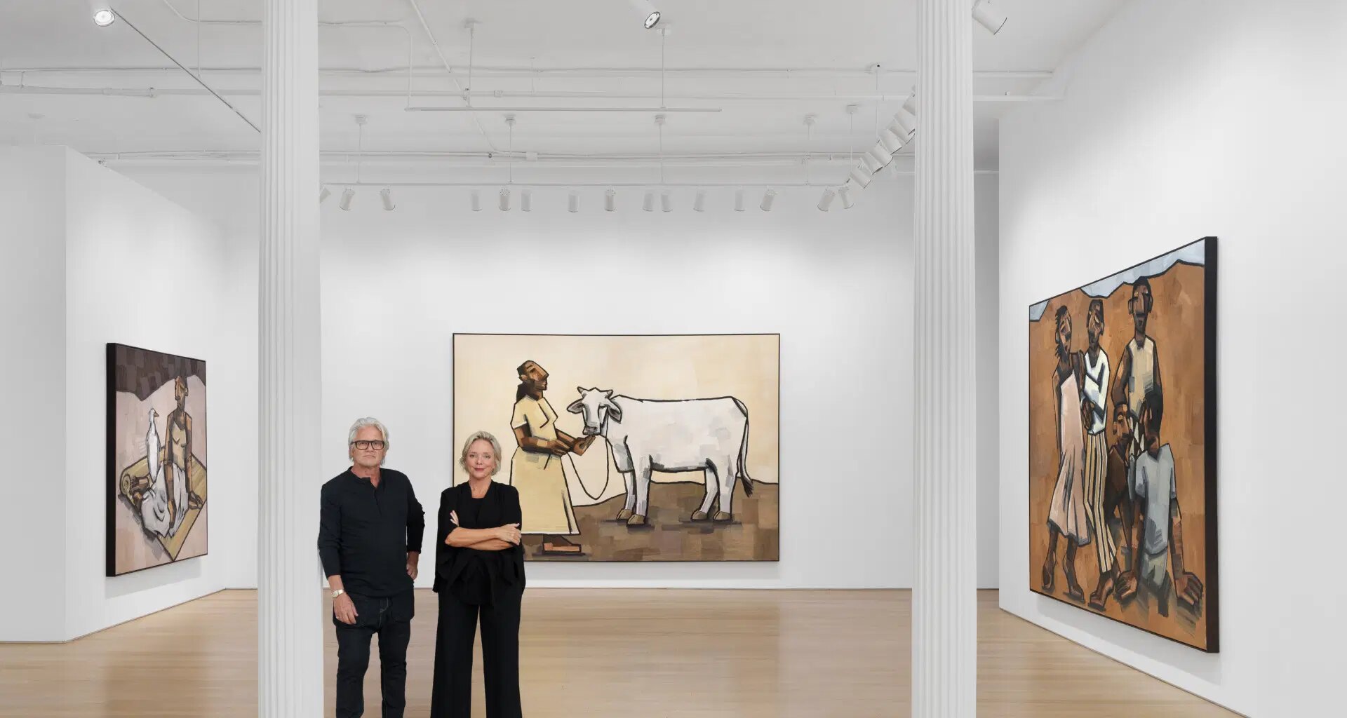 Two people standing in an art gallery with large paintings depicting figures and a cow on the white walls.