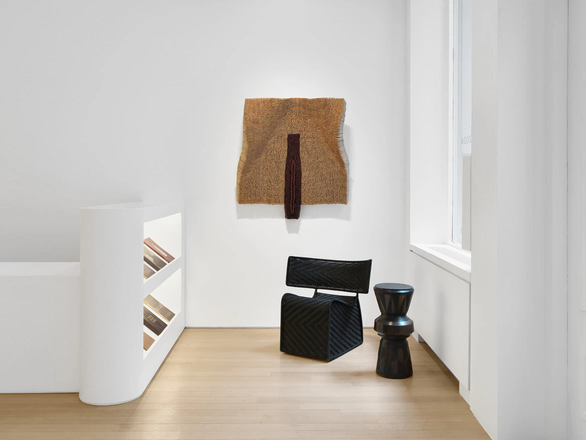 Modern art exhibit with woven textile on wall, black chair, and wooden stool in a bright, minimalist gallery space.