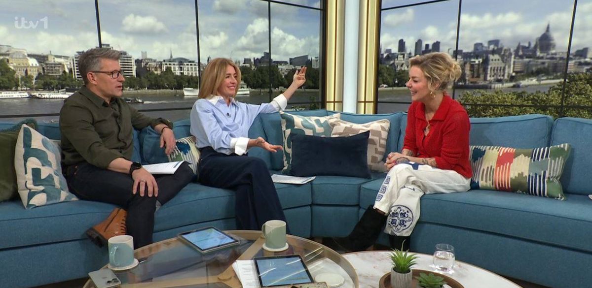Three individuals are engaged in a conversation on a talk show, seated on a blue couch, with a coffee table in front of them. The setting features large glass windows offering a view of a cityscape, and various decorative items adorn the couch and table.