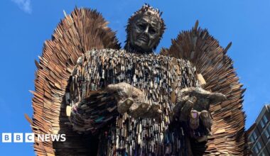 A tall male figure towers above a bright blue sky. The statues hands are open and shoulders are shrugged. The statue is made out of rusty knife blades of all different shapes and sizes.