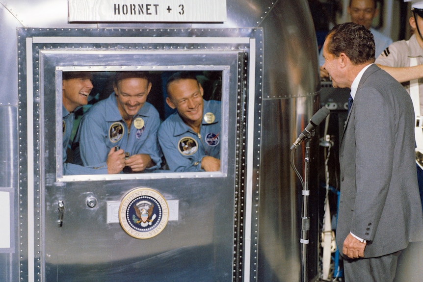 1970s photo showing a silver box with a window in it; three smiling men are looking out while a man in a suit looks in.