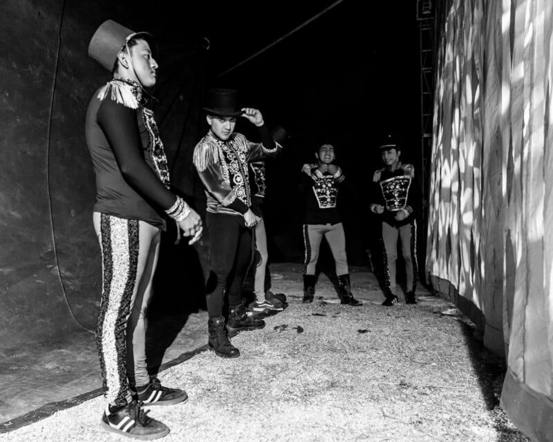 Five male circus performers wearing decorative costumes and top hats stand backstage under a tent. Some are adjusting their outfits, while others wait, with dramatic lighting and shadows cast on the tent wall.