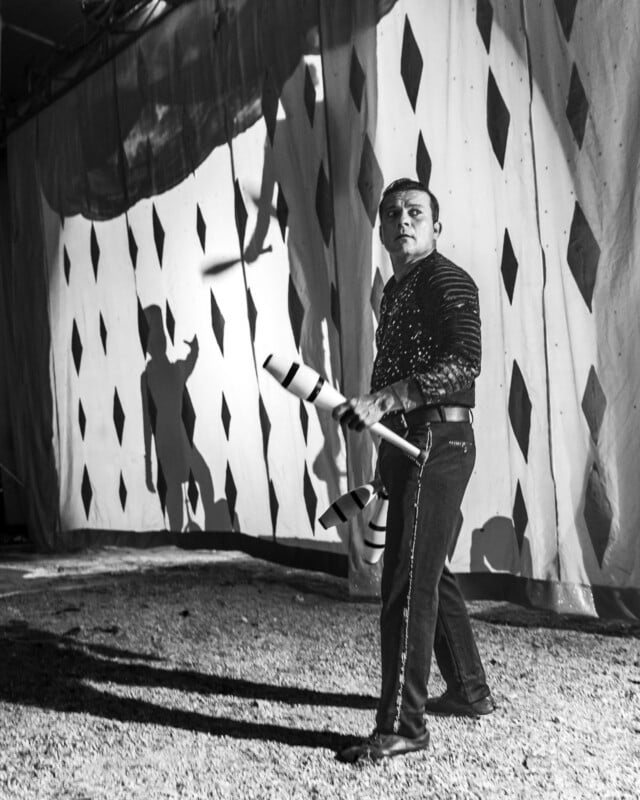 A man in a sequined shirt juggles clubs in front of a diamond-patterned curtain, casting dramatic shadows on the backdrop in a black-and-white circus setting.
