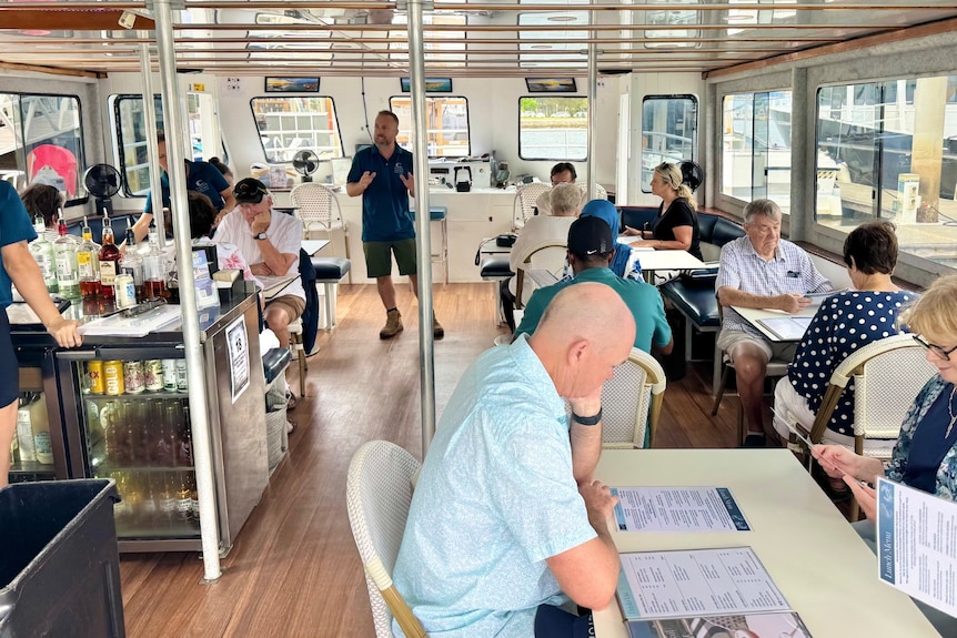 People sitting at tables and chairs on a boat, while a man in uniform stands and speaks.
