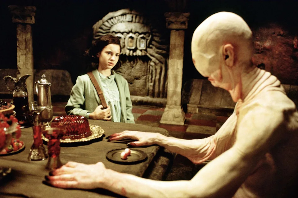 Scene from fantasy film with a young girl facing a pale, humanoid creature at a feast table in an underground setting